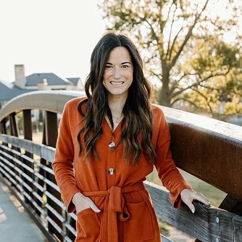 Whitney Martin standing on a bridge wearing a orange coat with her hand in her pocket. She is smiling  